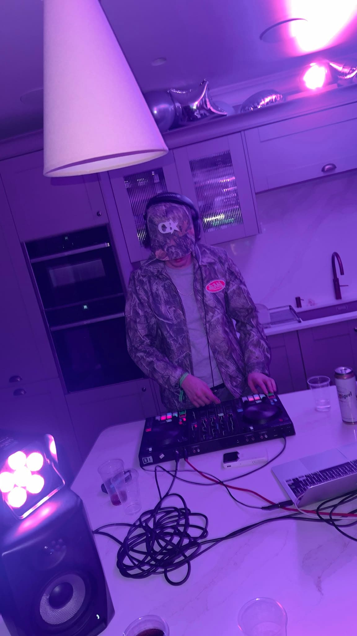 DJ KOZZ performing in a camo jacket and cap on a compact DJ controller under purple kitchen lighting