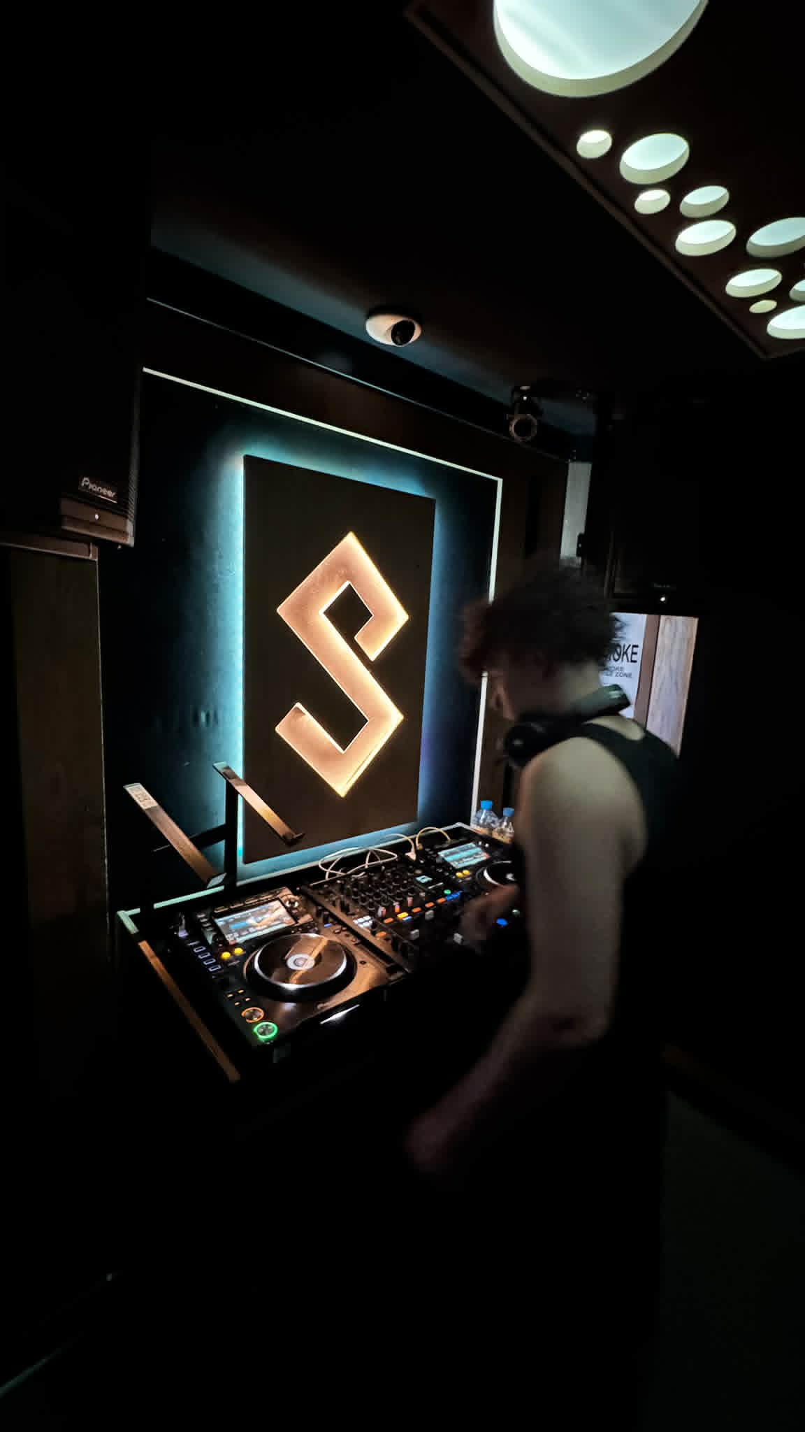 DJ KOZZ at Pioneer CDJ setup with cyan S logo backdrop