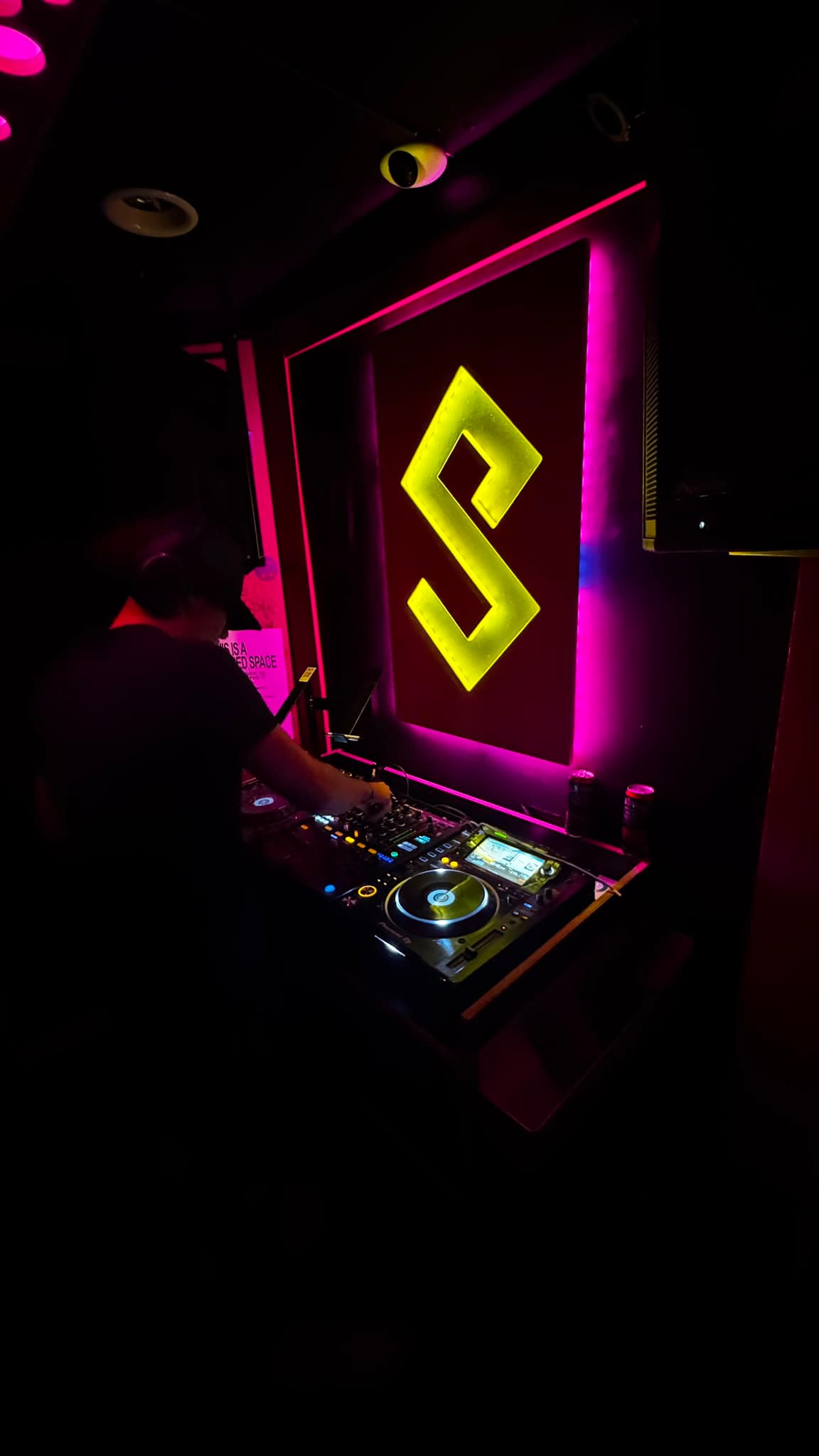 DJ KOZZ silhouetted on Pioneer CDJs against a gold-lit illuminated S logo backdrop