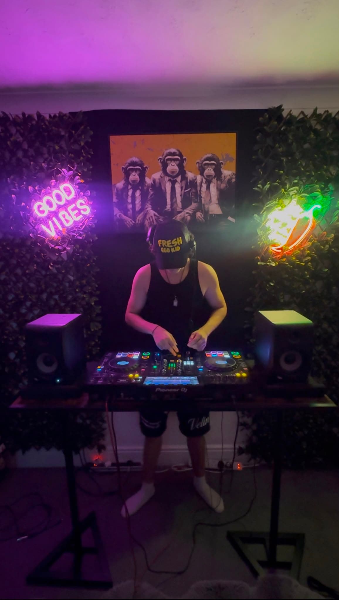 DJ KOZZ performing under a Good Vibes neon sign and three wise monkeys artwork, hedge backdrop