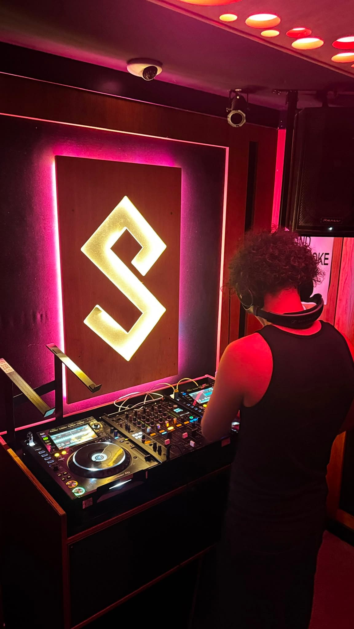 DJ KOZZ from behind on Pioneer CDJs, magenta-lit S logo backdrop and ceiling downlights