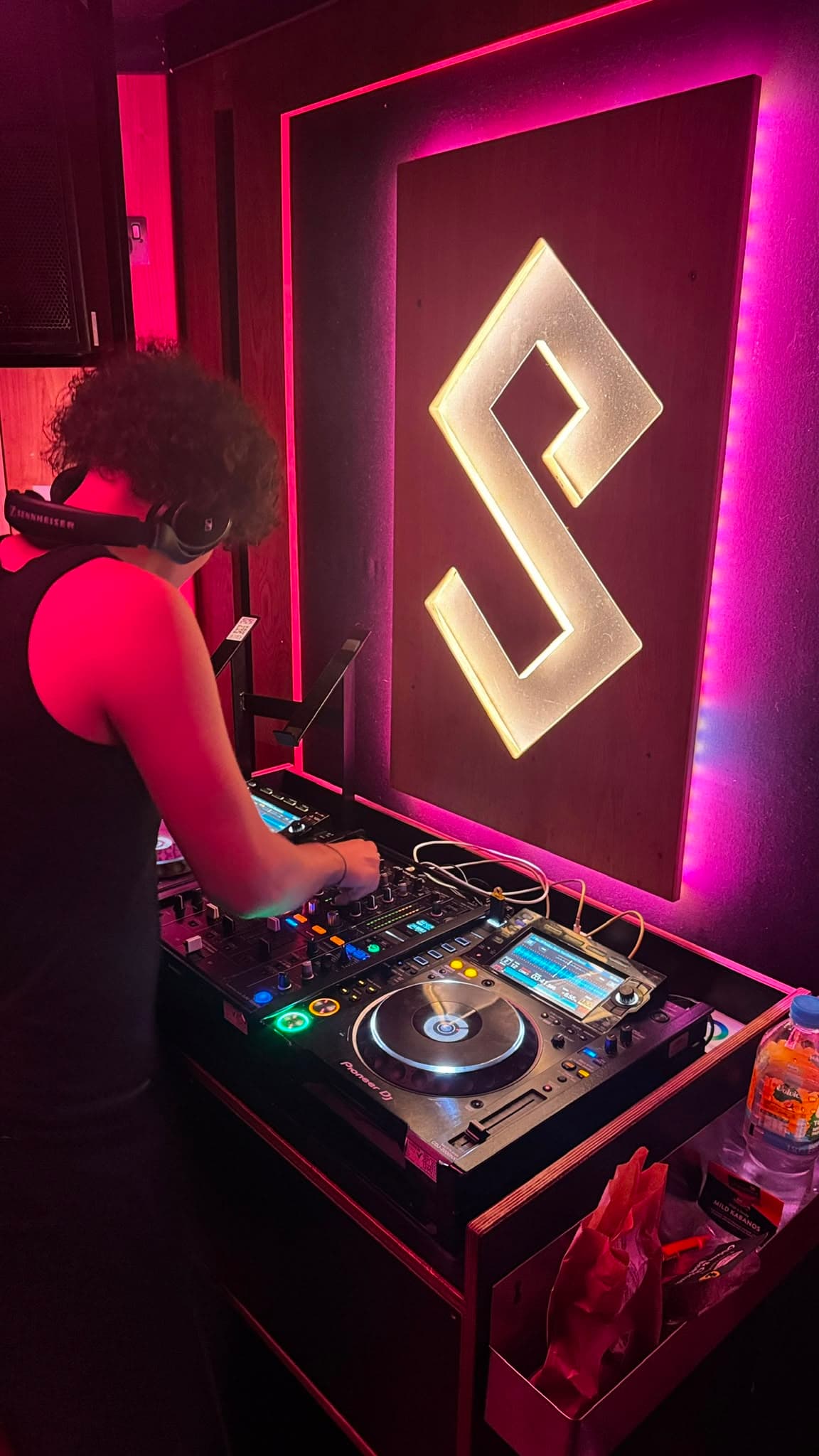 DJ KOZZ close-up on Pioneer CDJs against a magenta-lit illuminated S logo backdrop